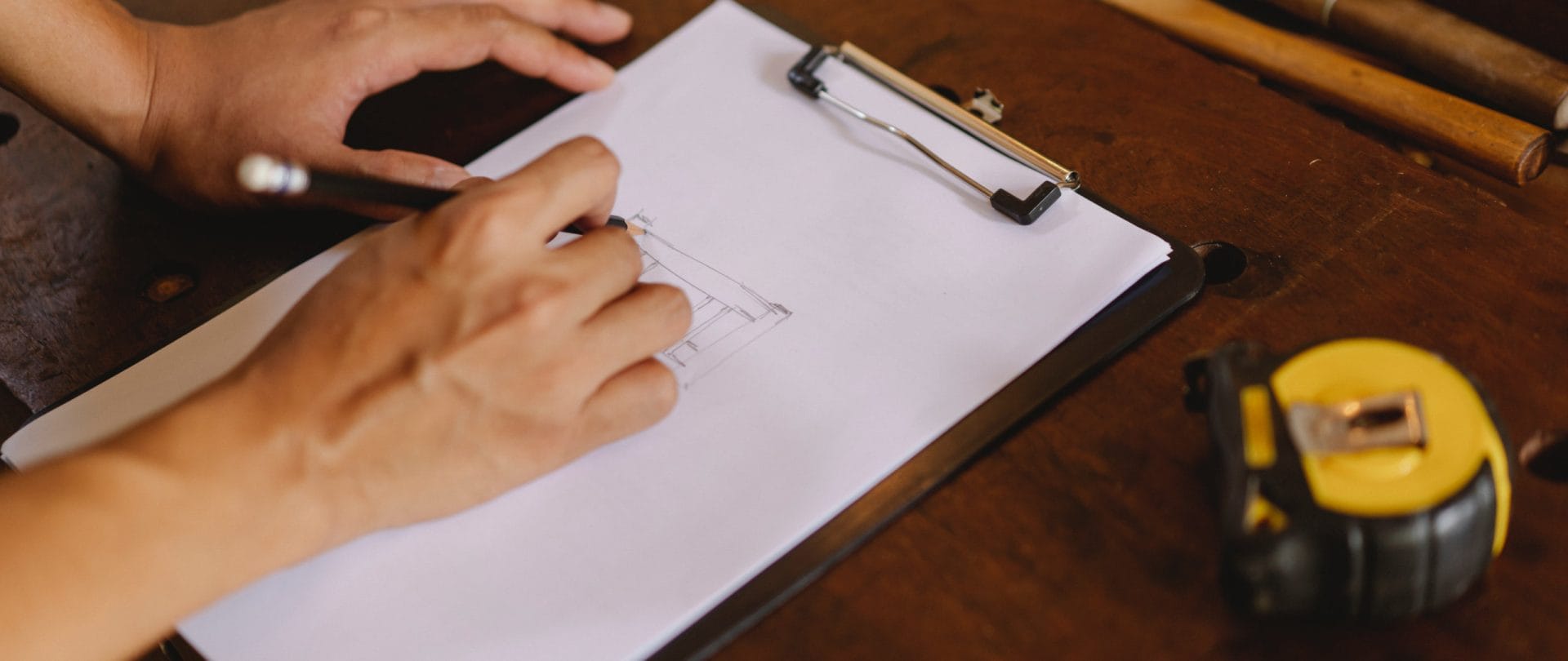 En person skitserer noget på et stykke papir, der er fastgjort til en klemmeplade. Klemmepladen ligger på en træbænk.