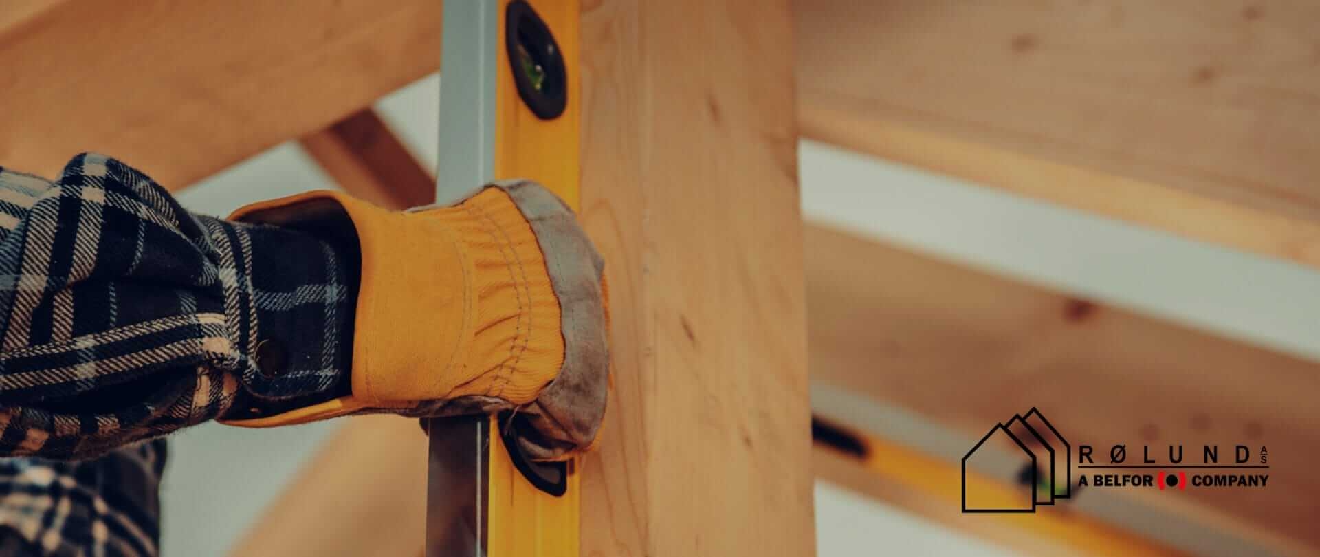 Et vaterpas holdes mod en træbjælke, personen bærer en gul arbejdshandske og en blå/hvid ternet skjorte.