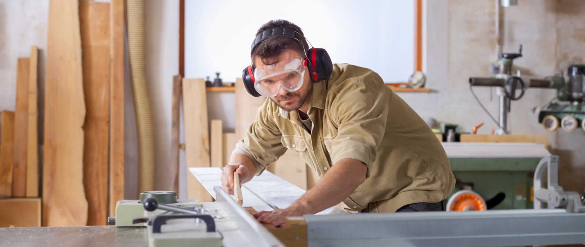 Mand i beskyttelsesdragt skubber træplade gennem en maskine